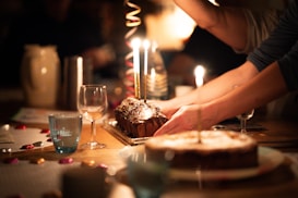 A warmly lit setting with a table displaying a chocolate loaf cake adorned with lit candles, surrounded by colorful candies and empty glassware. Hands are reaching in to adjust or present the cake, suggesting a celebratory occasion. The soft focus and ambient lighting create a cozy atmosphere.