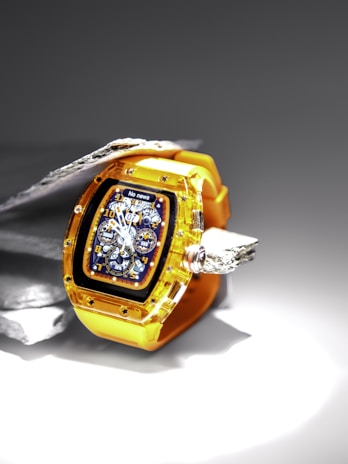 A luxury watch featuring a transparent yellow strap and casing, showcasing intricate gears and dials on a dark, detailed face. The watch is displayed against a contrasting background of metallic and stone textures, highlighting its craftsmanship.