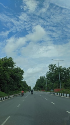 Several motorcyclists are traveling on a wide, open road flanked by green trees. The sky is partly cloudy with patches of blue visible.