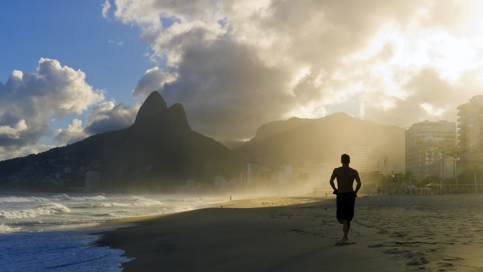 Homem correndo na areia da praia ao amanhecer, com o morro Dois Irmãos e os prédios da orla do Rio de Janeiro ao fundo