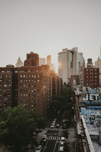 Urban setting featuring tall buildings with a mixture of modern glass skyscrapers and old brick structures. The street below is lined with trees and cars, with some graffiti visible on a rooftop. The sun is setting, casting a warm glow over the scene.