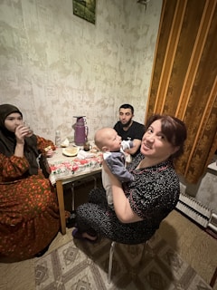 A cozy indoor scene with three adults and one baby gathered around a small table. The table is set with a floral tablecloth, and a purple thermos and some food items are visible. One woman is holding the baby and smiling at the camera. Another woman is drinking from a glass, and a man is seated in the background. The room features patterned wallpaper and a curtain with warm tones.