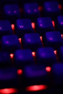 A close-up view of a keyboard with red backlighting, highlighting keys with a soft purple glow. The focus is on the Q, W, E, R, and D keys, set against a dark background, creating a vivid contrast.