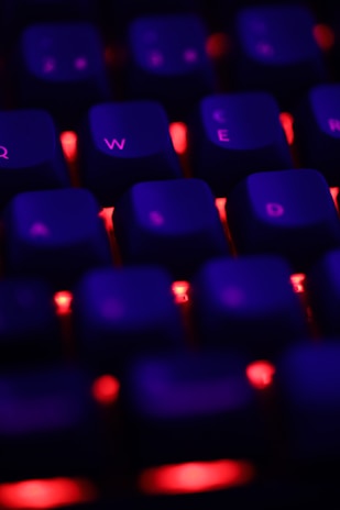 A close-up view of a keyboard with red backlighting, highlighting keys with a soft purple glow. The focus is on the Q, W, E, R, and D keys, set against a dark background, creating a vivid contrast.