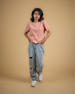 A model wearing a casual white t-shirt and jeans standing against a soft pink wall.