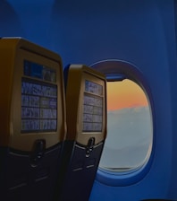 Interior of an airplane showing two seat-back screens and a window with a view of a colorful sky transitioning from blue to orange, suggesting a sunrise or sunset.