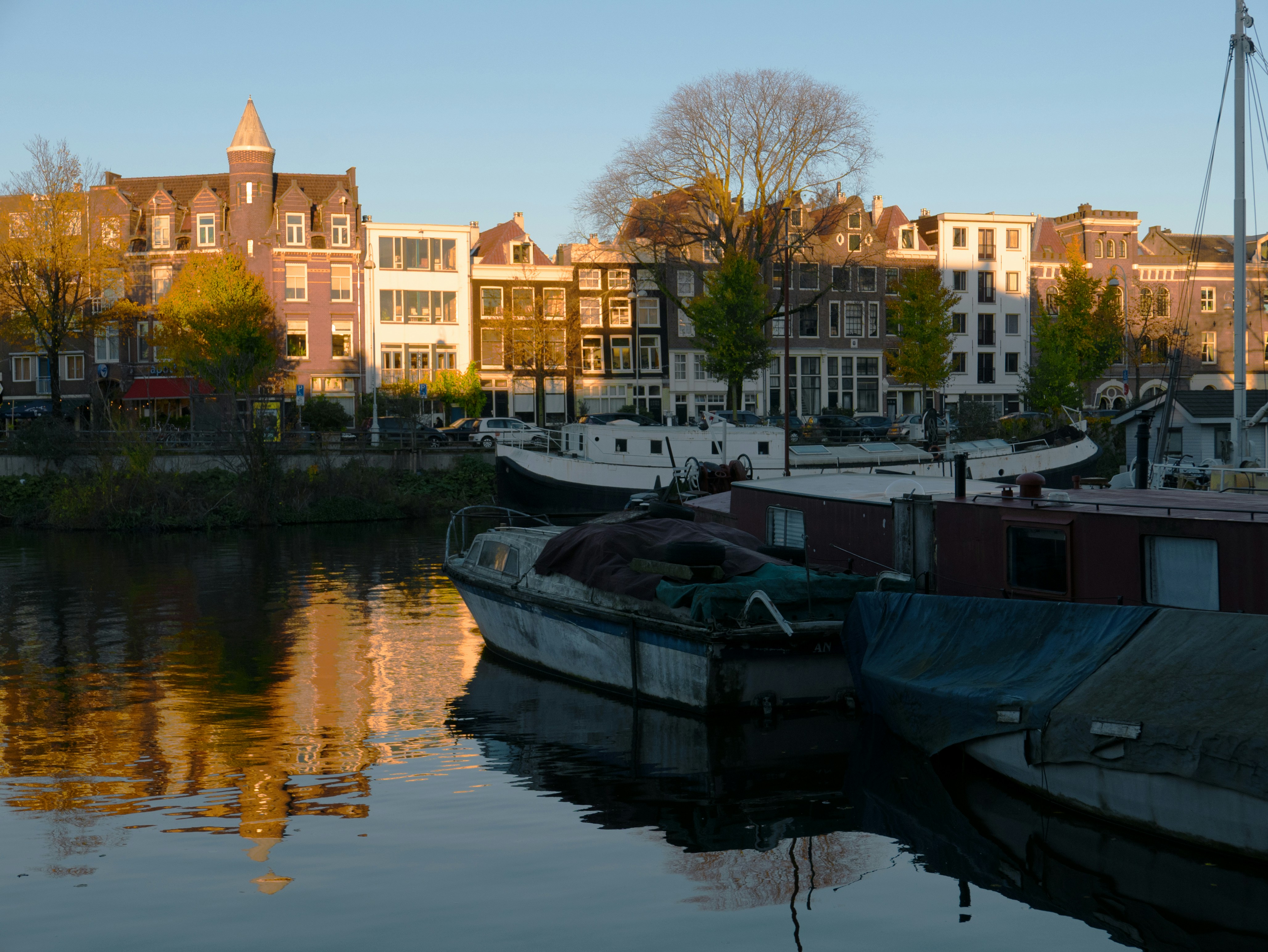 a row of houses next to a body of water, A canal view in the city Amsterdam with sunset light and dark shadows. Boats are drifting in the reflecting water. Many light textures and shades. Street photography of old cities in The Netherlands by Fons Heijnsbroek, 2023 // Uitzicht met late zon over het stille water en de woonboten in de Nieuwe Vaart, met de huizen en bomen aan de Kattenburgergracht. Op een herfstdag met veel schaduw en blauw water - in Amsterdam stad, 2023. Gratis foto, Fons Heijnsbroek: fotografie straat in Nederland in hoge resolutie.
