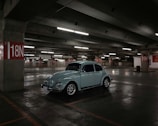 A vintage car is parked in a spacious, dimly lit underground parking garage. The concrete structure features red and white parking signs indicating various sections such as 18N and 19N. Overhead fluorescent lights cast soft illumination over the area, creating shadows and reflections on the polished concrete floor.