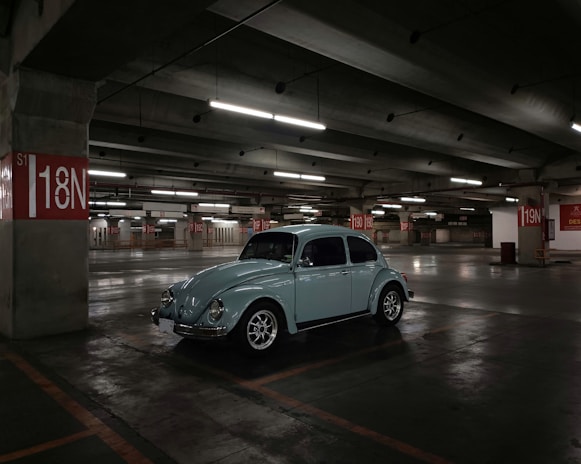 A vintage car is parked in a spacious, dimly lit underground parking garage. The concrete structure features red and white parking signs indicating various sections such as 18N and 19N. Overhead fluorescent lights cast soft illumination over the area, creating shadows and reflections on the polished concrete floor.