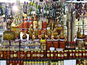 A vibrant display of various pickled and preserved foods in jars of different sizes. The shelves are packed with tightly arranged bottles and jars containing olives, peppers, and other vegetables. Above, hanging rows of olive oil bottles and condiments add to the busy, colorful scene. The setting appears to be a market or specialty store.