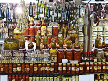 A vibrant display of various pickled and preserved foods in jars of different sizes. The shelves are packed with tightly arranged bottles and jars containing olives, peppers, and other vegetables. Above, hanging rows of olive oil bottles and condiments add to the busy, colorful scene. The setting appears to be a market or specialty store.