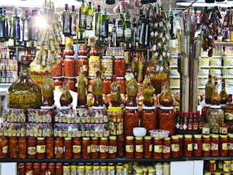 A vibrant display of various pickled and preserved foods in jars of different sizes. The shelves are packed with tightly arranged bottles and jars containing olives, peppers, and other vegetables. Above, hanging rows of olive oil bottles and condiments add to the busy, colorful scene. The setting appears to be a market or specialty store.