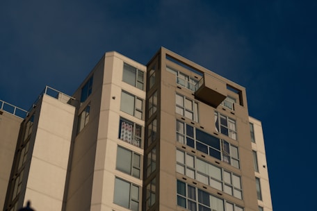 A modern high-rise apartment building under a clear blue sky, featuring large reflective windows and several balconies. The sunlight is casting soft shadows on the facade.