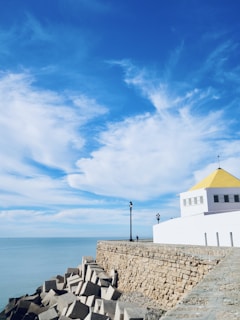 A coastal scene with a clear blue sky featuring a few scattered white clouds. A stone seawall runs along the shore, leading to a structure with a yellow roof. Several large geometric concrete blocks are placed at the edge of the water. A lamppost is positioned along the walkway atop the wall, and a person is seen walking in the distance.