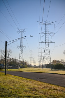 Several large metal transmission towers stand in a grassy area adjacent to a paved road, with numerous power lines stretching across the clear, blue sky. In the background, more towers continue in a line, creating a sense of depth and perspective. The morning sunlight casts soft shadows on the grass.