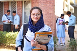 A young woman wearing glasses and a navy scarf stands in the foreground, holding textbooks labeled with accounting terms. She is dressed in a light blue uniform. In the background, a group of students are engaged in activities such as reading books and discussing among themselves, set against a brick building.