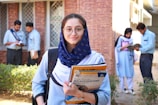 A young woman wearing glasses and a navy scarf stands in the foreground, holding textbooks labeled with accounting terms. She is dressed in a light blue uniform. In the background, a group of students are engaged in activities such as reading books and discussing among themselves, set against a brick building.