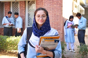 A young woman wearing glasses and a navy scarf stands in the foreground, holding textbooks labeled with accounting terms. She is dressed in a light blue uniform. In the background, a group of students are engaged in activities such as reading books and discussing among themselves, set against a brick building.