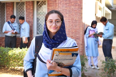A young woman wearing glasses and a navy scarf stands in the foreground, holding textbooks labeled with accounting terms. She is dressed in a light blue uniform. In the background, a group of students are engaged in activities such as reading books and discussing among themselves, set against a brick building.