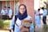 A young woman wearing glasses and a navy scarf stands in the foreground, holding textbooks labeled with accounting terms. She is dressed in a light blue uniform. In the background, a group of students are engaged in activities such as reading books and discussing among themselves, set against a brick building.