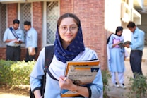 A young woman wearing glasses and a navy scarf stands in the foreground, holding textbooks labeled with accounting terms. She is dressed in a light blue uniform. In the background, a group of students are engaged in activities such as reading books and discussing among themselves, set against a brick building.