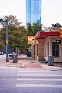 A street view with a crosswalk leading towards a building with stone facade and a red roof. Above the entrance, there is a sign for 'Gus's World Famous Fried Chicken'. In the background, there are trees and a tall modern glass building.