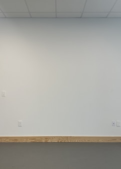 A plain, light-colored wall with a wooden baseboard near the bottom. The wall has electrical outlets and a light switch. The ceiling features a grid-like pattern with panels.