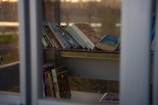 A collection of books is displayed on a shelf inside a glass enclosure. The books are varied in size and color, and a few are leaning against each other, suggesting they may have been recently placed. The enclosure appears to be a small library or book exchange, with glass panels allowing natural light to filter in. The overall scene is quiet and serene, suggesting an inviting place for reading.