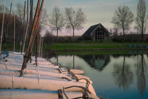 A serene lakeside scene features a row of small sailboats docked along the shore in the foreground. Behind them, a modern A-frame cabin with large windows stands surrounded by trees. The calm water reflects the cabin and trees, enhancing the tranquil atmosphere.