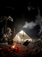 A cozy snow camping setup with a glowing tent surrounded by snow-covered trees at dusk.