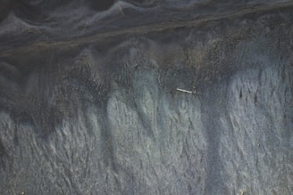 Dark sand with flowing, natural patterns resembling liquid or waves, possibly due to water or wind erosion.