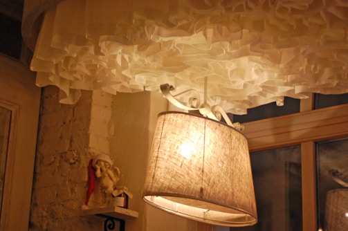 A warmly lit room featuring a large fabric ceiling lamp with a textured lampshade. The ceiling is adorned with ruffled white fabric panels. A small cherub figurine with a red Santa hat is sitting on a wall-mounted shelf against a textured, rustic brick wall.