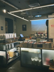 A cozy bookstore with shelves lined with books, a counter with a computer, and various items like coffee machines on the back counter. The space has industrial elements with exposed piping and pendant lights, and a projection screen displaying an image on the wall.