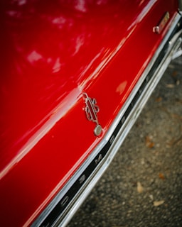 A shiny red car hood with chrome details and a decorative emblem near the edge. The surface reflects light, indicating a well-polished and clean appearance. The background appears to be an asphalt surface with some scattered leaves.