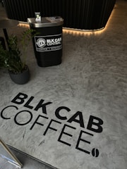 A modern interior with a textured floor featuring the words 'BLK CAB COFFEE' in black lettering. A small counter labeled 'BLK CAB CENTRAL COFFEE + KITCHEN' with promotional material and a plant next to it. Soft ambient lighting highlights a dark-colored wall.