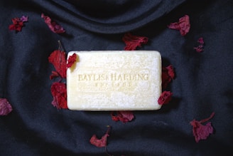 A bar of soap imprinted with 'Baylis & Harding England' placed on a black fabric background, surrounded by scattered red petals.