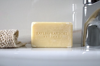 A bar of Baylis & Harding soap is placed upright on a bathroom counter. Beside it lies a rolled-up beige textured cloth on the left, and a shiny metal faucet is visible on the right. The background is a smooth, glossy white surface reflecting light.