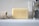 A bar of Baylis & Harding soap is placed upright on a bathroom counter. Beside it lies a rolled-up beige textured cloth on the left, and a shiny metal faucet is visible on the right. The background is a smooth, glossy white surface reflecting light.