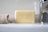 A bar of Baylis & Harding soap is placed upright on a bathroom counter. Beside it lies a rolled-up beige textured cloth on the left, and a shiny metal faucet is visible on the right. The background is a smooth, glossy white surface reflecting light.