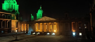 A historical building illuminated at night with green and warm orange lights highlighting domes and columns. The architecture features classical elements with a symmetrical design, ornate carvings, and a prominent pediment.