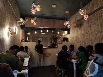 A cozy café interior with various people seated at tables, engaging in conversation. The ambiance features decorative pendant lights hanging from the ceiling, walls adorned with textured wallpaper, and a colorful menu displayed above the counter. A mix of single and group seating is available, and there is a warm, inviting atmosphere.
