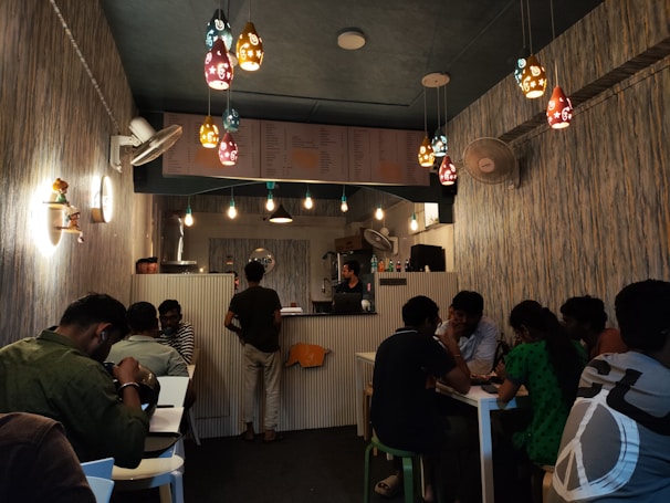 A cozy café interior with various people seated at tables, engaging in conversation. The ambiance features decorative pendant lights hanging from the ceiling, walls adorned with textured wallpaper, and a colorful menu displayed above the counter. A mix of single and group seating is available, and there is a warm, inviting atmosphere.