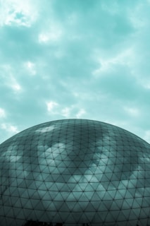 A large geodesic dome with a reflective surface is shown, capturing the cloudy sky above it. The structure consists of numerous triangular panels, creating a pattern across its surface. The sky is overcast with a soft, teal tint.