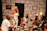 A group of five people are sitting and standing in a room with a stone-patterned wall. They are engaged in discussion or storytelling, with expressions of amusement and interest. The room has a few decorative elements, including a wall clock, a painting, and a yellow lamp on a side table. The individuals are dressed in a variety of stylish outfits, including a white dress, a light blue dress, and a light suit with a hat.