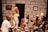 A group of five people are sitting and standing in a room with a stone-patterned wall. They are engaged in discussion or storytelling, with expressions of amusement and interest. The room has a few decorative elements, including a wall clock, a painting, and a yellow lamp on a side table. The individuals are dressed in a variety of stylish outfits, including a white dress, a light blue dress, and a light suit with a hat.