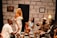 A group of five people are sitting and standing in a room with a stone-patterned wall. They are engaged in discussion or storytelling, with expressions of amusement and interest. The room has a few decorative elements, including a wall clock, a painting, and a yellow lamp on a side table. The individuals are dressed in a variety of stylish outfits, including a white dress, a light blue dress, and a light suit with a hat.