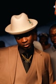 A well-dressed individual wearing a tan pinstripe suit, black shirt, and layered gold necklaces. The person dons a stylish cream fedora with a decorative pin, standing under warm lighting with others in the background.