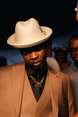 A well-dressed individual wearing a tan pinstripe suit, black shirt, and layered gold necklaces. The person dons a stylish cream fedora with a decorative pin, standing under warm lighting with others in the background.