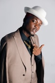 A person dressed in a pinstriped suit and black shirt, accessorized with gold jewelry and rings. They are wearing a stylish white hat tilted to one side. The person is striking a confident pose against a plain background.