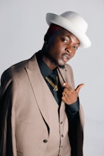 A person dressed in a pinstriped suit and black shirt, accessorized with gold jewelry and rings. They are wearing a stylish white hat tilted to one side. The person is striking a confident pose against a plain background.